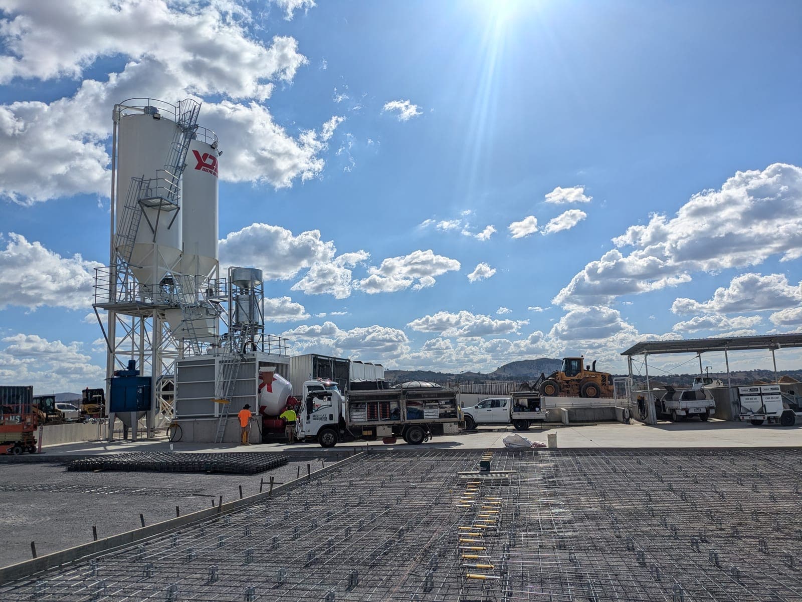 A concrete batching plant with a tall white silo and elevated hopper, seen across a wide expanse of reinforcing mesh laid ready for a slab pour
