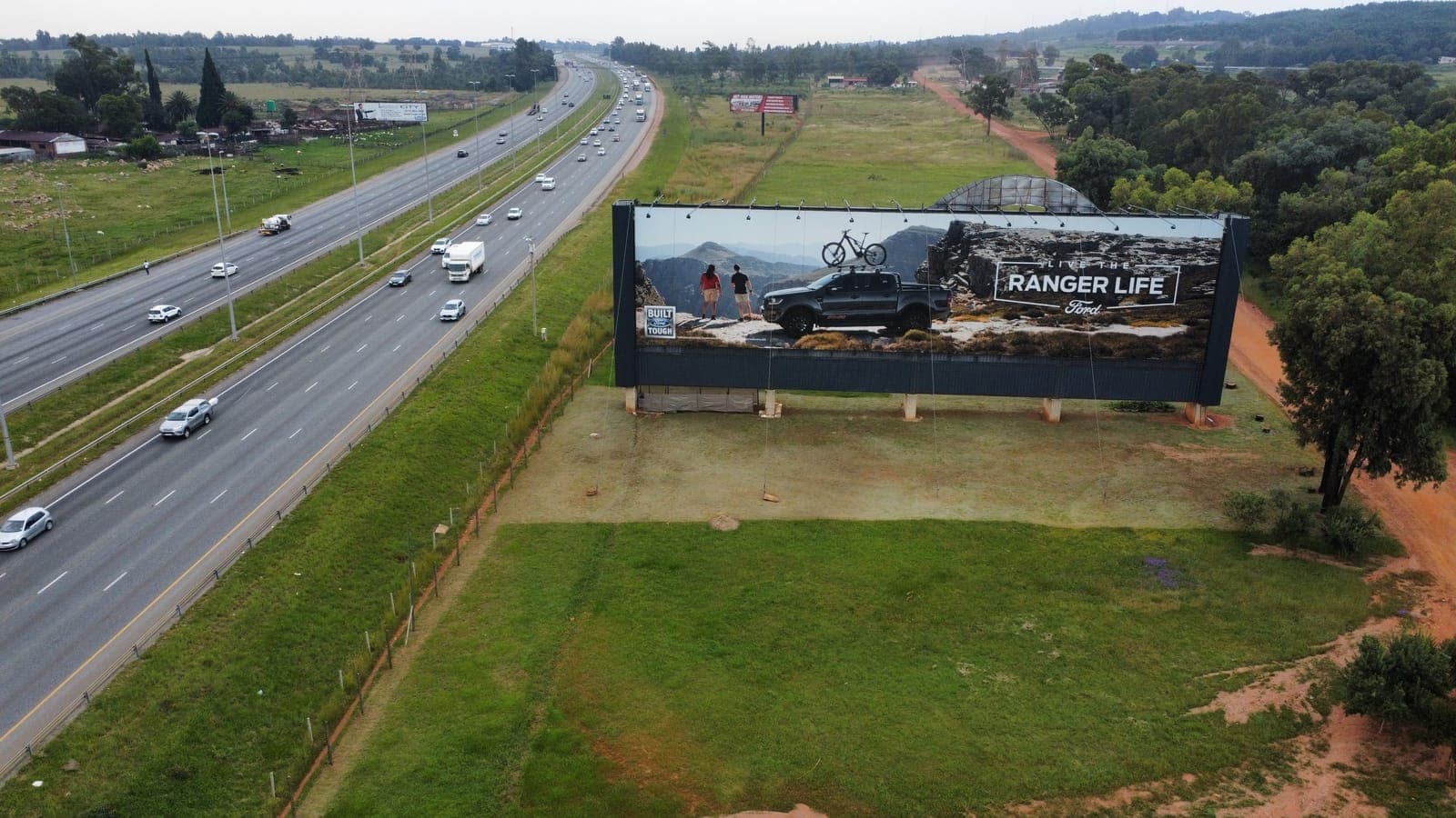 Aerial view of a large static billboard on a steel support frame standing in open ground beside a divided highway