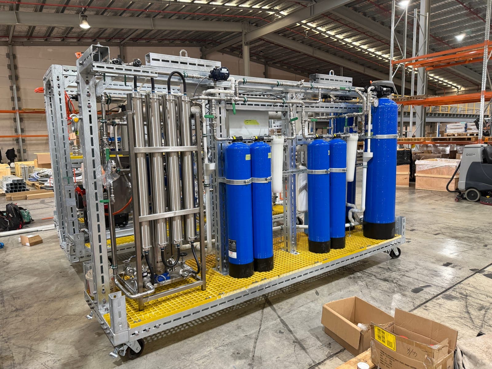 A skid-mounted treatment plant built into a galvanised steel frame, with blue pressure vessels and stainless pipework, standing on a warehouse floor