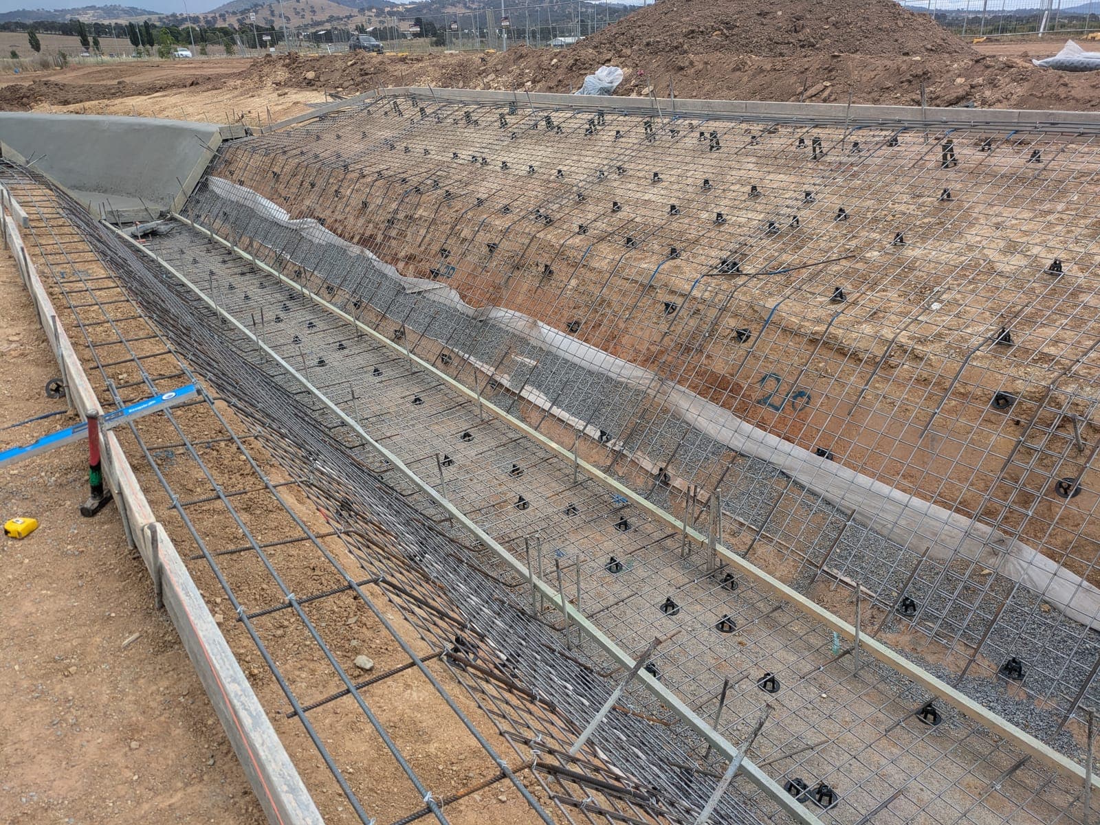 A long retaining wall under construction, its cut batter faced with steel reinforcing mesh and anchor plates above a reinforced footing