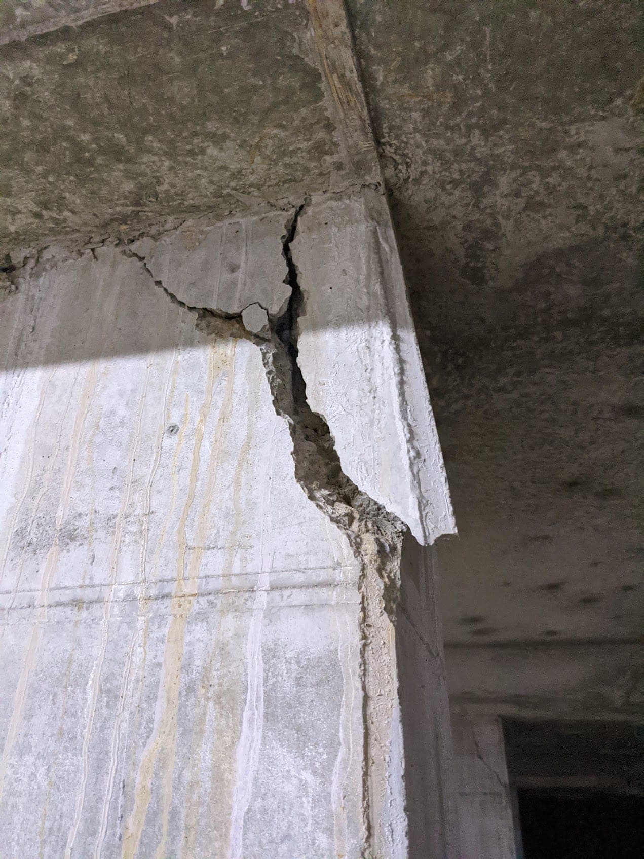 A deep crack splitting a concrete beam where it meets the slab soffit, the concrete spalled away along the crack and pale mineral staining running down the wall below