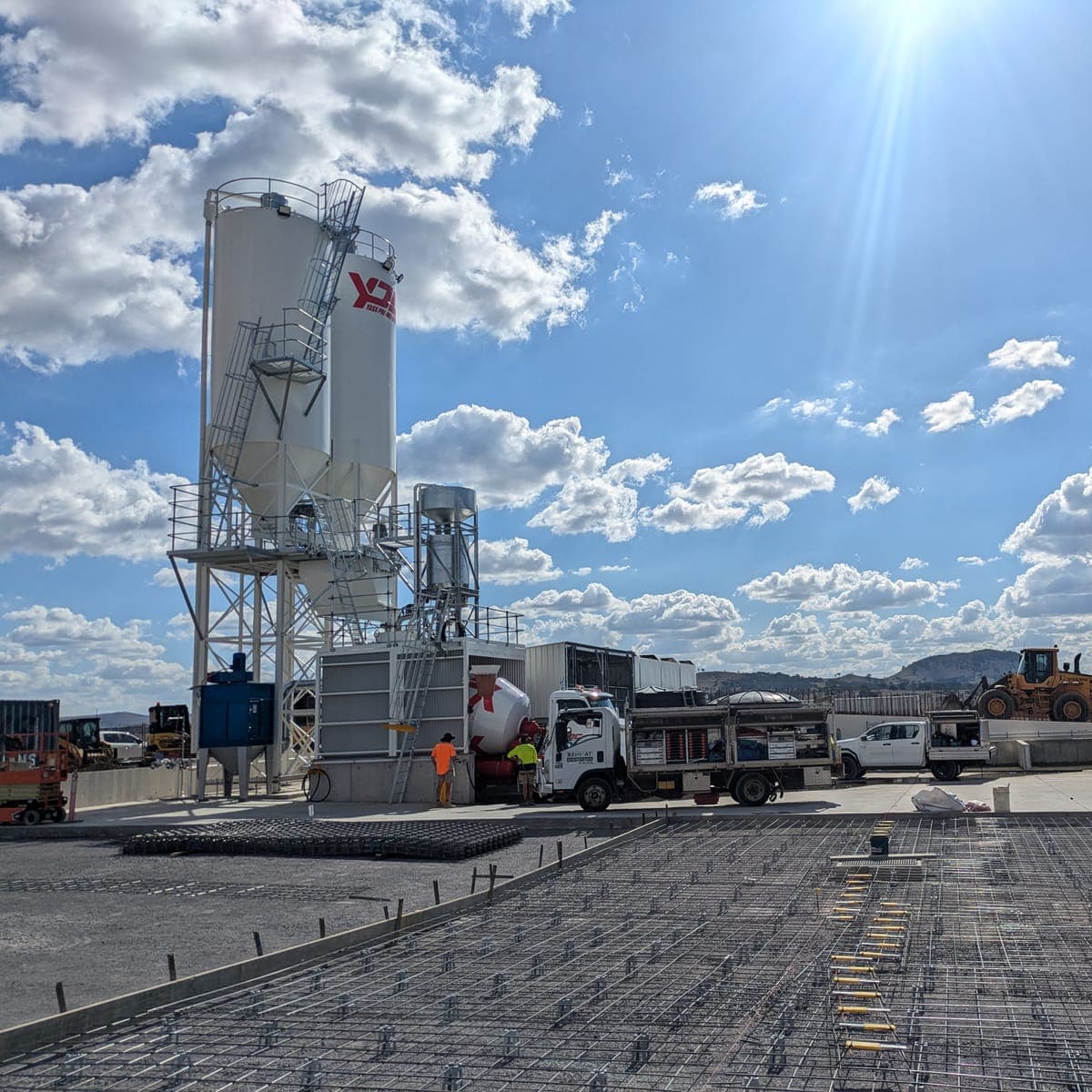 A concrete batching plant on an open site, its tall white silo carried on a steel support frame with access ladders and walkways, an agitator truck and a front-end loader alongside, and reinforcing mesh laid across the slab in the foreground