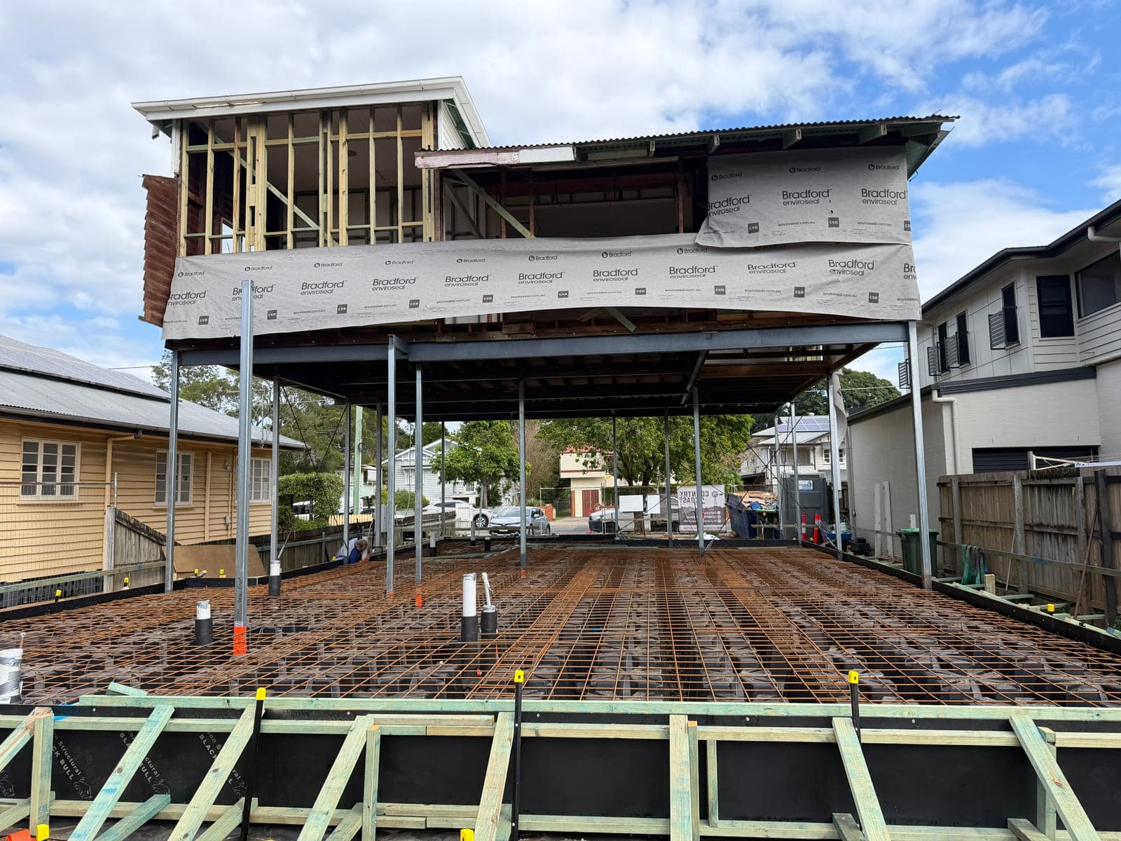 A weatherboard house raised on steel posts above newly laid reinforcing mesh and formwork for a ground-floor slab
