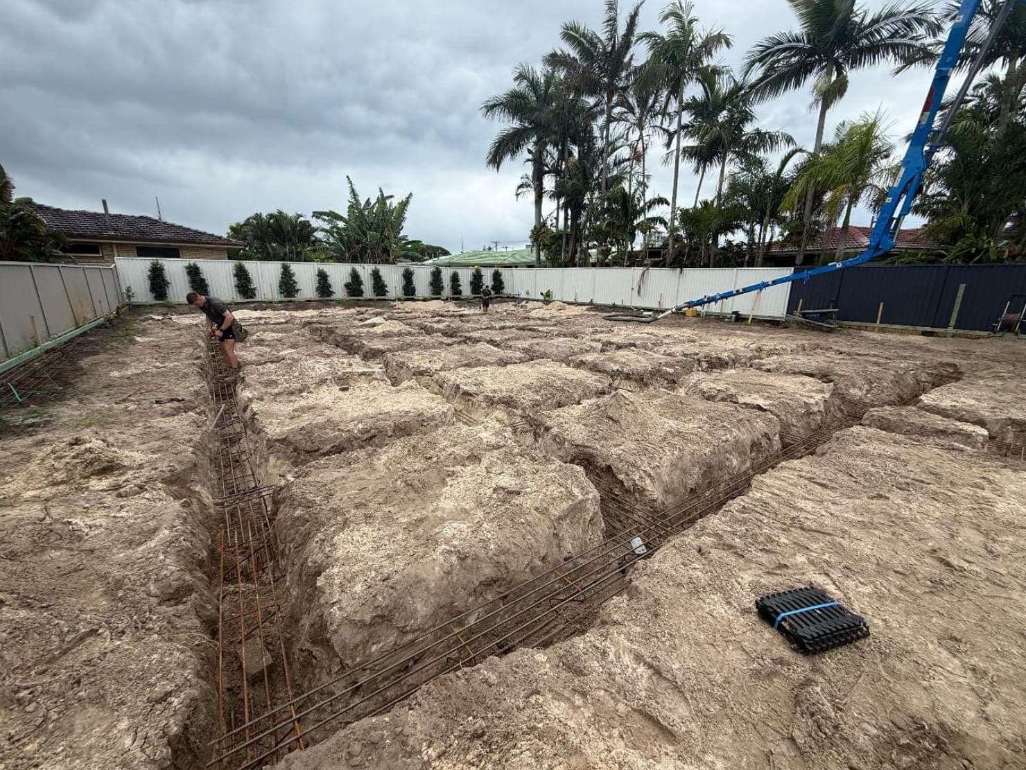 Excavated footing trenches across a sandy site with steel reinforcement laid in them, and a concrete pump boom reaching overhead