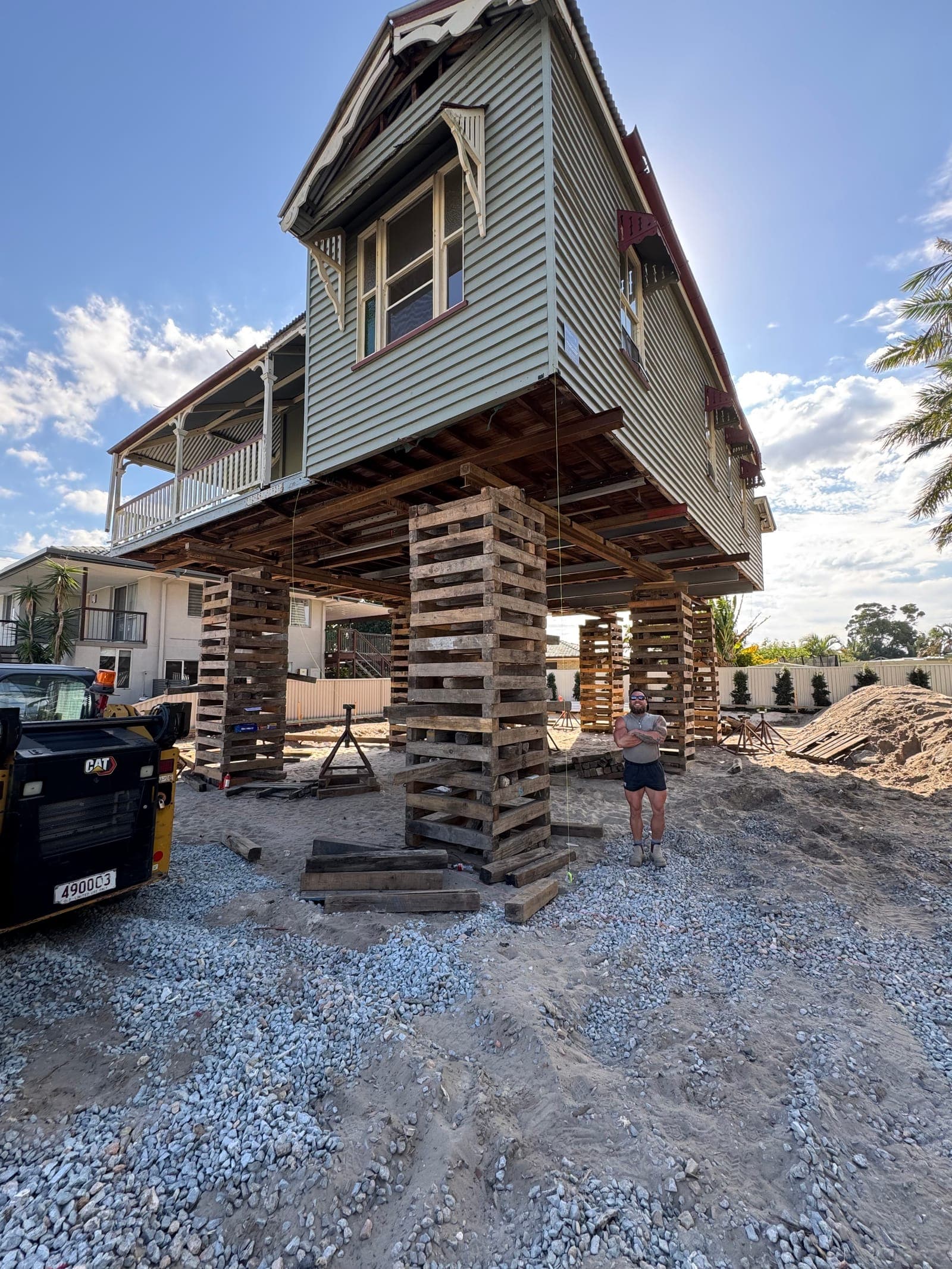 The house held high on stacked timber cribbing towers above a sand pad, with a person standing beneath it for scale