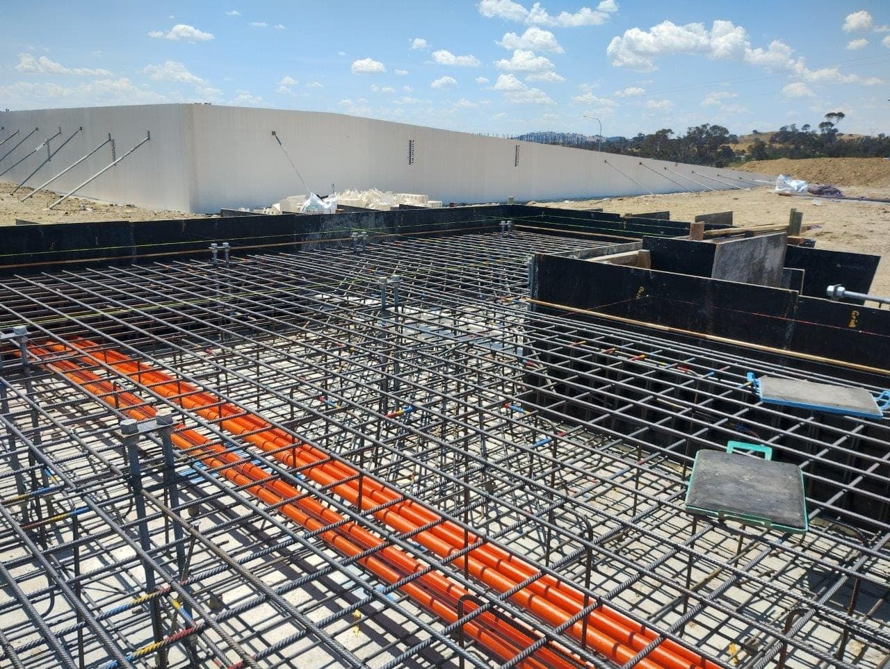 Densely tied steel reinforcement and orange service conduits laid within timber and steel formwork, with precast concrete panels standing beyond